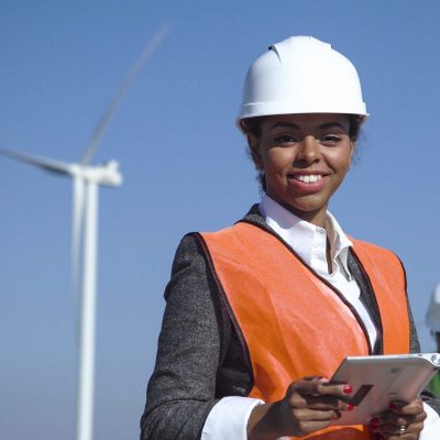 Female engineer wearing hardhat standing with digital tablet against other engineers and wind turbine on sunny day and looking at camera